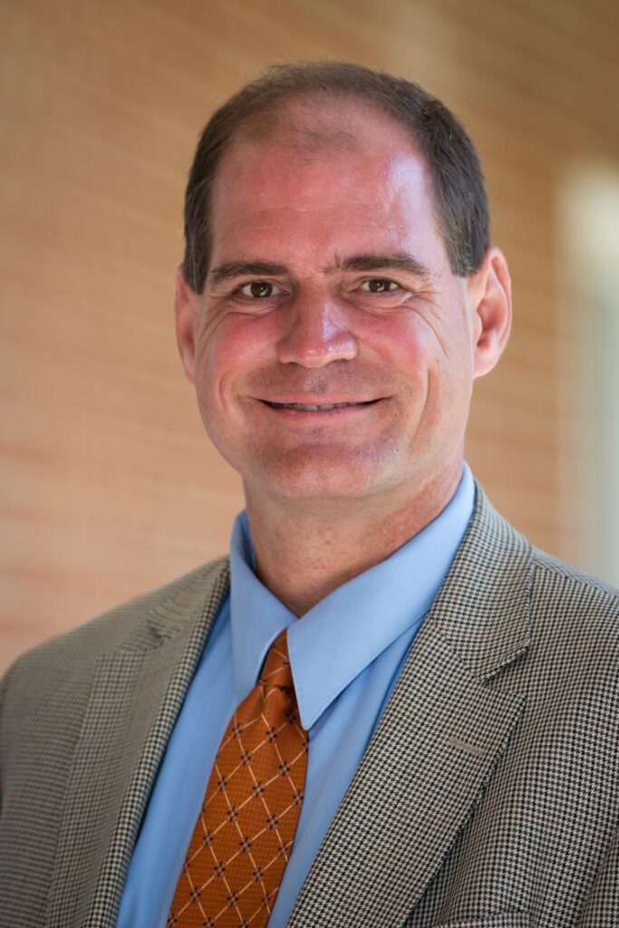 Portrait of Ian Danielsen, Assistant Professor at Longwood University. Ian is wearing a blue button up shirt with an orange tie and a plaid blazer.